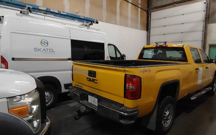 A yellow GMC four-by-four pickup parked beside a ladder-racked SKATEL van inside the shop.